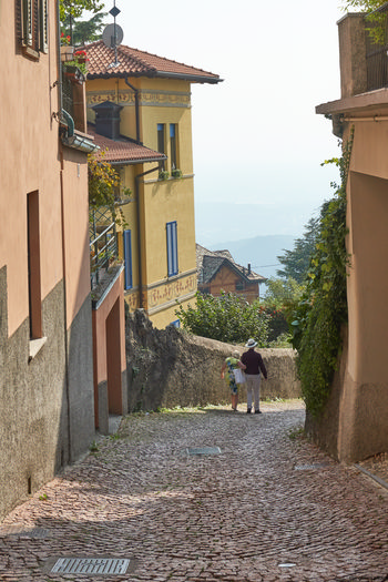 Brunate street This urban photograph captures a cobbled street in Brunate, a small town near Como, Italy, during the afternoon in late summer. The main subject of the image is the street itself, lined with pastel-colored architecture typical of the region, including a prominent yellow building with blue shutters and decorative trim. Pedestrians are visible in the foreground, specifically Jamie Weinbren and Patricia Weinbren, walking together down the gently curving street. The perspective also reveals a glimpse of the scenic landscape in the distance, while lush greenery and shrubs adorn the facades, contributing to the characteristic charm of northern Italian towns.